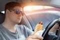 Adult male caucasian person holding hot dog fast food snack while driving car on sunny day. Men eating food while driving steering