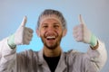 An adult male dressed in a white lab coat with a hair net on. He is smiling and giving a thumbs up. He is ready to work in a clean