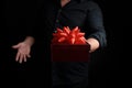 Adult man in a black shirt holds a red square box with a knotted bow on a dark background Royalty Free Stock Photo