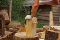 Adult man in gloves preparing firewood for campfire, holding log and chopping wood using ax, standing against house. Royalty Free Stock Photo