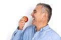 Adult man with grey hair wearing blue shirt ready to bite red applestanding on white background, dietology and healthy