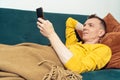 Adult man lie on sofa and watch video on smartphone in room. Person reclining on pillow under blanket and lounging.