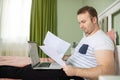 Adult man lying in his bed while he is working from home on his laptop. Papers or bills in his hand Royalty Free Stock Photo