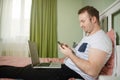 Adult man lying in his bed while he is working from home on his laptop. Phone in his hand