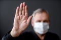 An adult man in a mask with his palm raised in protest on a dark background.