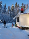 An adult man shovels snow from the roof of the veranda of a country house with a shovel, a young man removes snow below, a girl