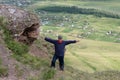 An adult man stands, arms outstretched, on a high cliff, against the backdrop of a countryside
