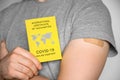 Adult man wearing gray t-shirt holding an International Certificate of Vaccination and shows his arm after vaccination