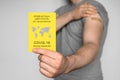 Adult man wearing gray t-shirt holding an International Certificate of Vaccination and shows his arm after vaccination
