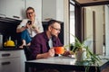 Adult married couple in home kitchen interior. Husband with beard sitting at table, looking at laptop, wife in glasses in the