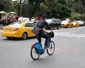 Adult member of the public seen riding a bicycle beside a moving New York taxi.