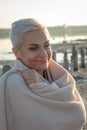 Adult middle-aged blonde woman wrapped herself in a bright plaid on the beach and looks at the sea, selective focus Royalty Free Stock Photo