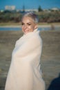 Adult middle-aged blonde woman wrapped herself in a bright plaid on the beach and looks at the sea, selective focus Royalty Free Stock Photo