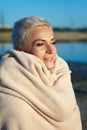 Adult middle-aged blonde woman wrapped herself in a bright plaid on the beach and looks at the sea, selective focus Royalty Free Stock Photo