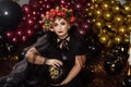 An adult plump woman in the image of a witch with flowers on her head sits in a dark studio Royalty Free Stock Photo