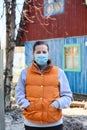 Adult Russian woman in safety mask stands against rural timber house, self-isolation in village or sparcely populated area