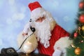 Adult santa claus in red suit sits at the table, calls on old phone, tree is beautifully decorated with balls, the concept of