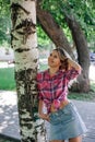Adult shining blonde woman with face looking upward standing in park leaning on birch wearing checkered shirt tied in