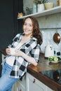 Adult smiling brunette woman in casual with cup of tea looking at camera on the kitchen