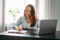 Adult smiling brunette woman doing notes in daily book with opened laptop. She study something with online course at the home Royalty Free Stock Photo