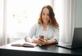 Adult smiling brunette woman doing notes in daily book and using the mobile phone with opened laptop at home Royalty Free Stock Photo