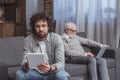 adult son using tablet and senior father using laptop on sofa