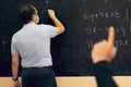 Adult teacher with mask writing on the blackboard with a student raising hand. Covid situation, pandemic, new normal