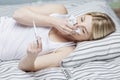 An adult white woman lying in bed in her pajamas checking her temperature on a thermometer and blowing her nose into a tissue. Royalty Free Stock Photo