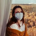 Adult woman in a medical mask by the window wrapped in a yellow plaid. Sad autumn weather and home comfort