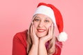 An adult woman in a Santa hat rests her chin with her hands on a pink background, close-up
