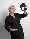 Adult woman with short hair, blonde. with glasses . She takes a selfie on the camera. There is a light background in the studio. C