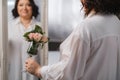 An adult woman in a white shirt is standing with a glass with roses sprinkled on it. A girl in the interior with a glass