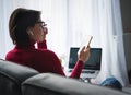 Adult woman working at home online holding phone video chatting with laptop large window in background in living room on sofa at