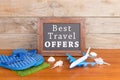 blackboard with text & x22;Best Travel OFFERS& x22;, plane, flops, seashells on brown wooden background