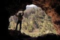 ADVENTURER EXPLORER WITH AUSTRALIAN HAT AND BACKPACK PUTTING HIS  HANDS THE ROOF OF A CAVE WATCHING A GREEN PINE FOREST Royalty Free Stock Photo