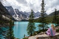 Adventurous blonde woman sits on the edge of a cliff along the rockpile trail at Moraine Lake, viewing the Canadian Rockies