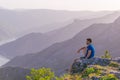 Adventurous man sitting on top of a mountain and enjoying the beautiful view, while looking downhill at the blue river and amazing