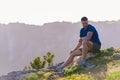 Adventurous man sitting on top of a mountain and enjoying the beautiful view, while looking downhill at the blue river and amazing