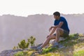 Adventurous man sitting on top of a mountain and enjoying the beautiful view, while looking downhill at the blue river and amazing