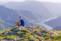 Adventurous man sitting on top of a mountain and enjoying the beautiful view, while looking downhill at the blue river and amazing