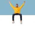 Advertising billboard template. Excited arab guy sitting on top of blank white poster with mockup, raising hands up
