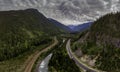 Aerial of cars on highway, train on railway tracks, river in between mountains on a cloudy day