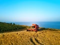 Aerial of red combine harvester working in wheat field near cliff with sea view on sunset. Harvesting machine cutting