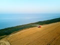 Aerial of red combine harvester working in wheat field near cliff with sea view on sunset. Harvesting machine cutting