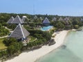 Aerial of resort bungalows and a pool, atop a short cliff and beside a white sand beach and tropical waters during a clear day.