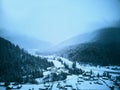 Aerial view of foggy countryside and houses in snowy valley. Hills and mountains with pine tree forest covered in snow