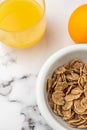 Aerial view of a glass of juice, a bowl of cereals and an orange on white marble, in vertical