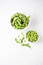 Aerial view of pods in drainer and edamame grains in white bowl, on white table