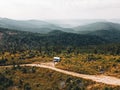 Aerial view of small camper trailer in the middle of summer forest, with campers sitting by fire.