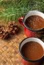 Aerial view of two red cups with chocolate, with falling sugar, on a table braided with branches and pine cones, vertically,
